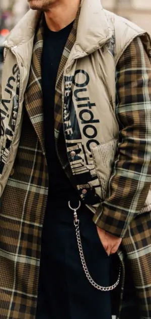 Men’s streetwear look with a plaid overcoat layered under an outdoor-themed vest.