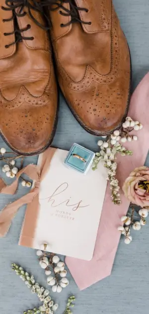Men's wedding suits ideas, An elegant flat lay of men’s wedding shoes, a vow book, a ring box, a pink tie, and florals.