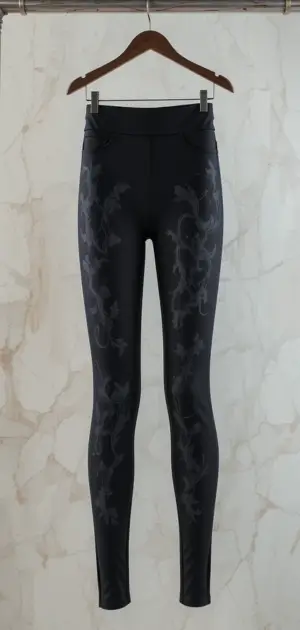 Here are the extracted details based on the image:**Description:** Sleek black office leggings with subtle floral detailing for a modern touch. **Caption:** Elegant black office leggings with a stylish floral embossed design. **Alt text:** A pair of black office leggings with floral patterns hanging on a wooden hanger.